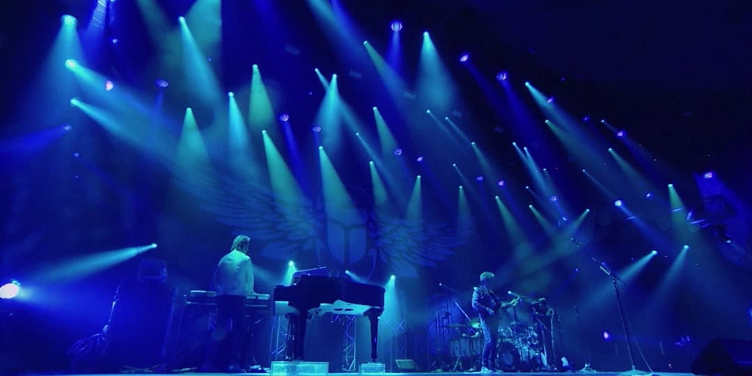 Journey performs on a stage with blue lighting and spotlights. The set includes a keyboard, a drum kit, and several guitars. A winged emblem is visible on the backdrop.