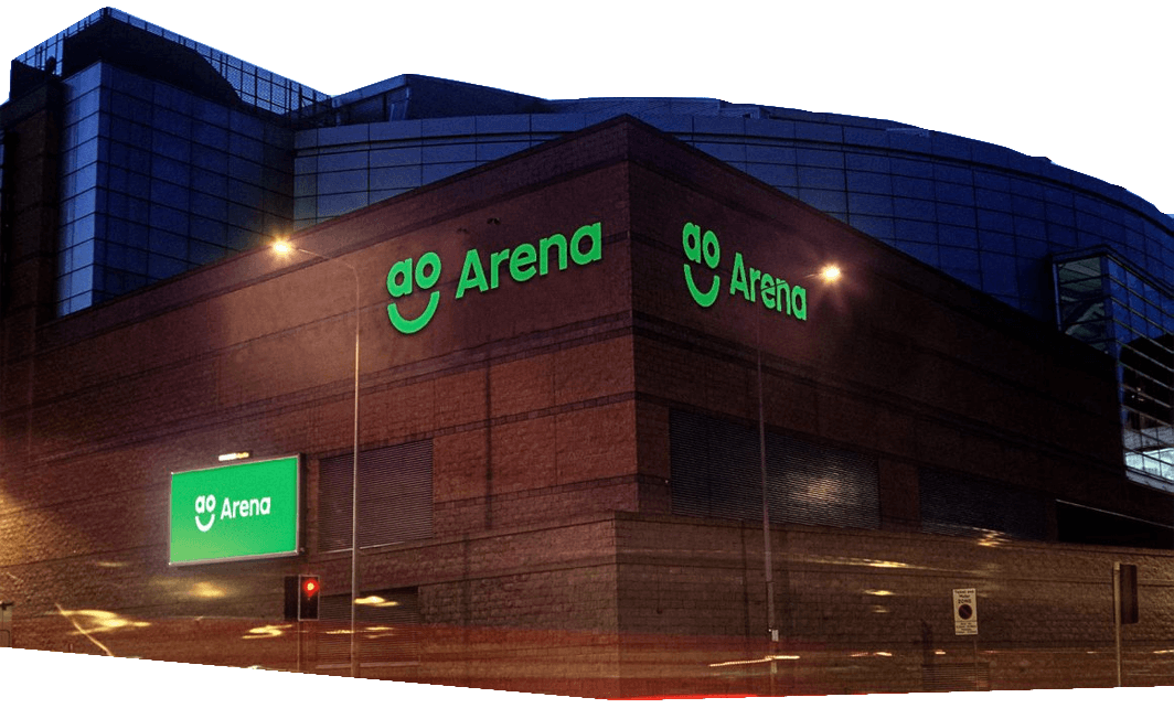 The exterior of AO Arena, a large brick and glass event venue, with green illuminated "AO Arena" signs and smiley face logos at night.