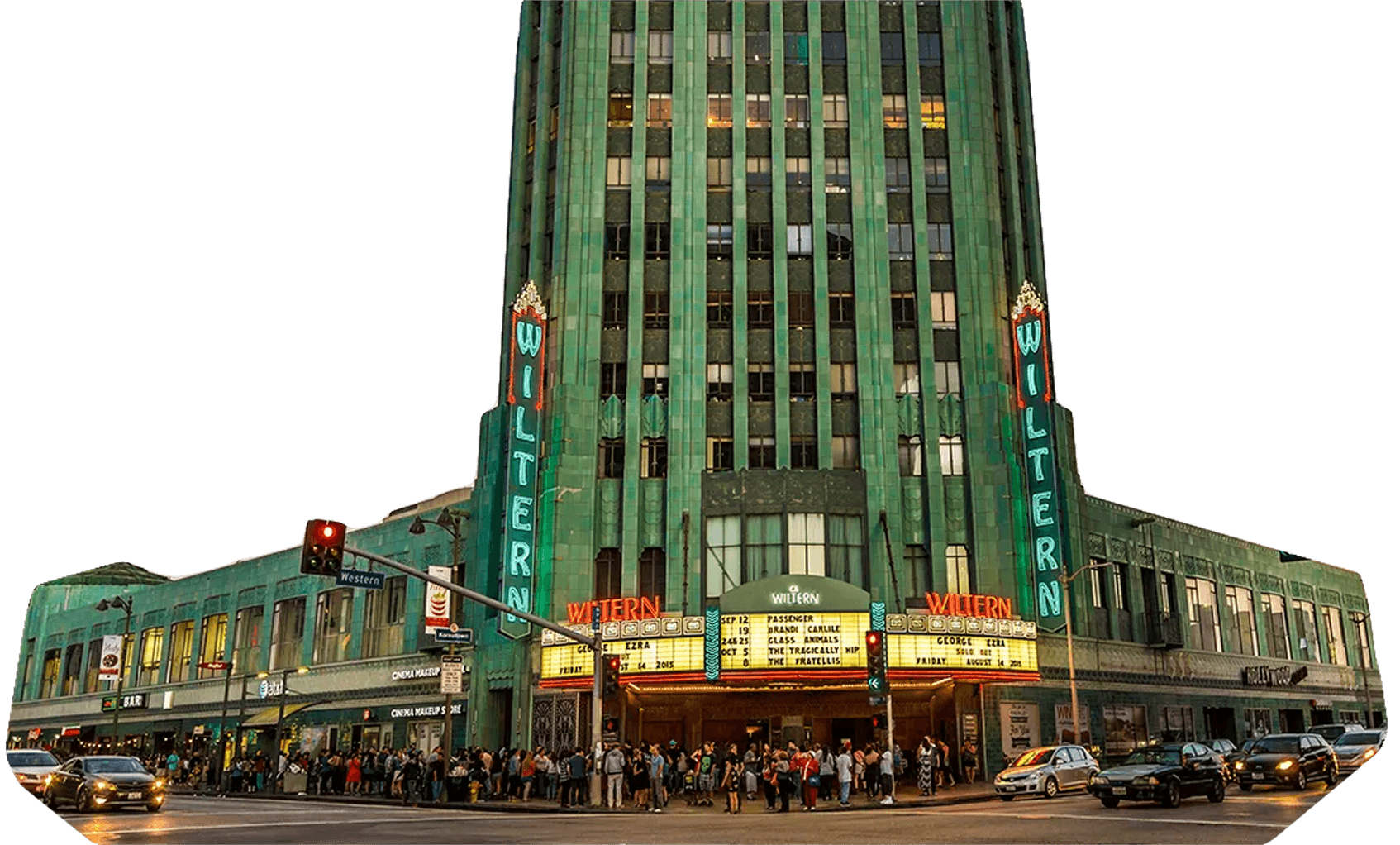 Art Deco Wiltern Theatre at dusk with green facade, neon signs, and a crowd lined up outside; cars and traffic lights visible in the foreground.