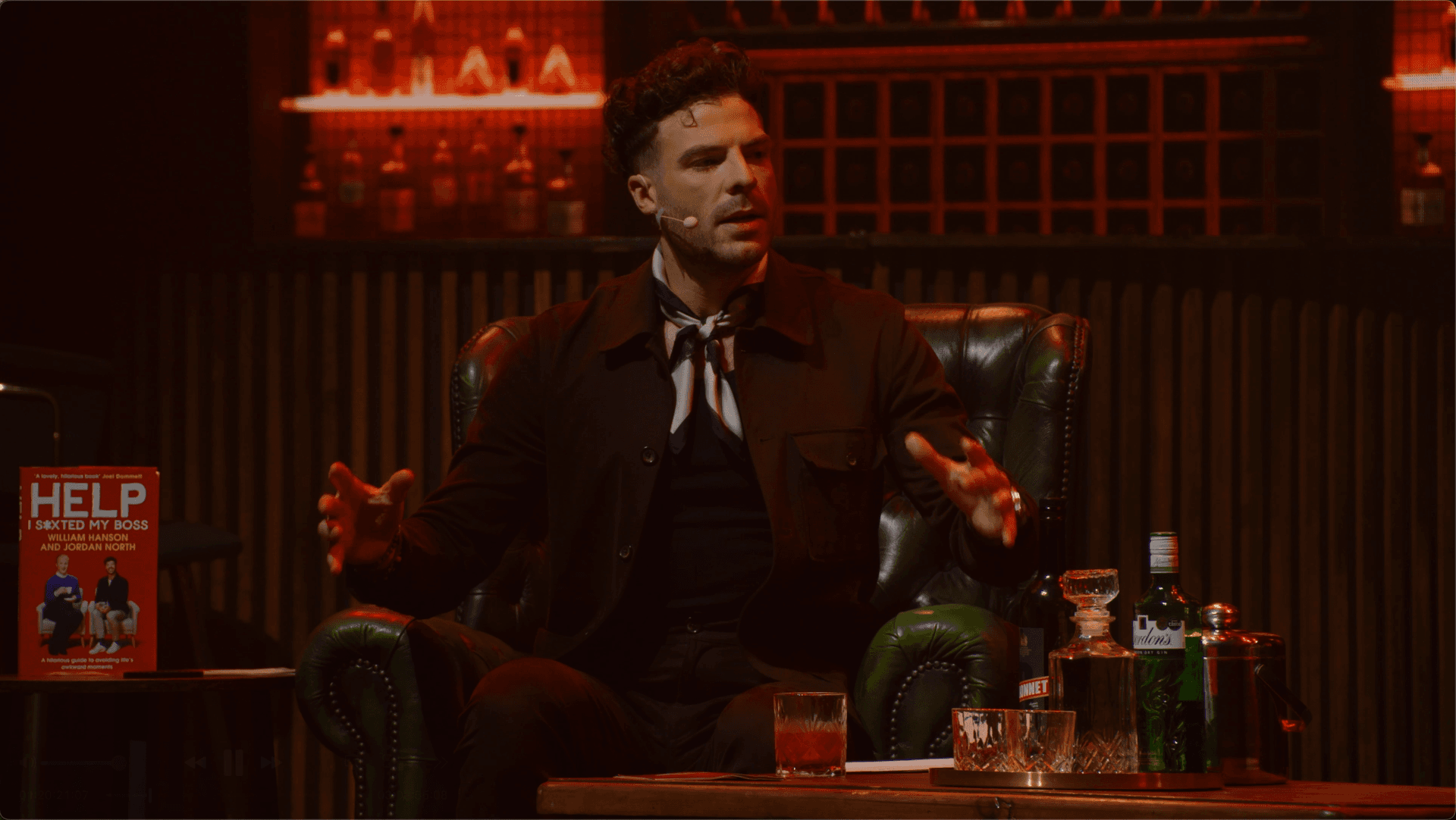 A man sits in a green leather armchair on stage, speaking with expressive hand gestures. A book, glasses, and bottles are on the table beside him. Shelves with bottles are in the background.