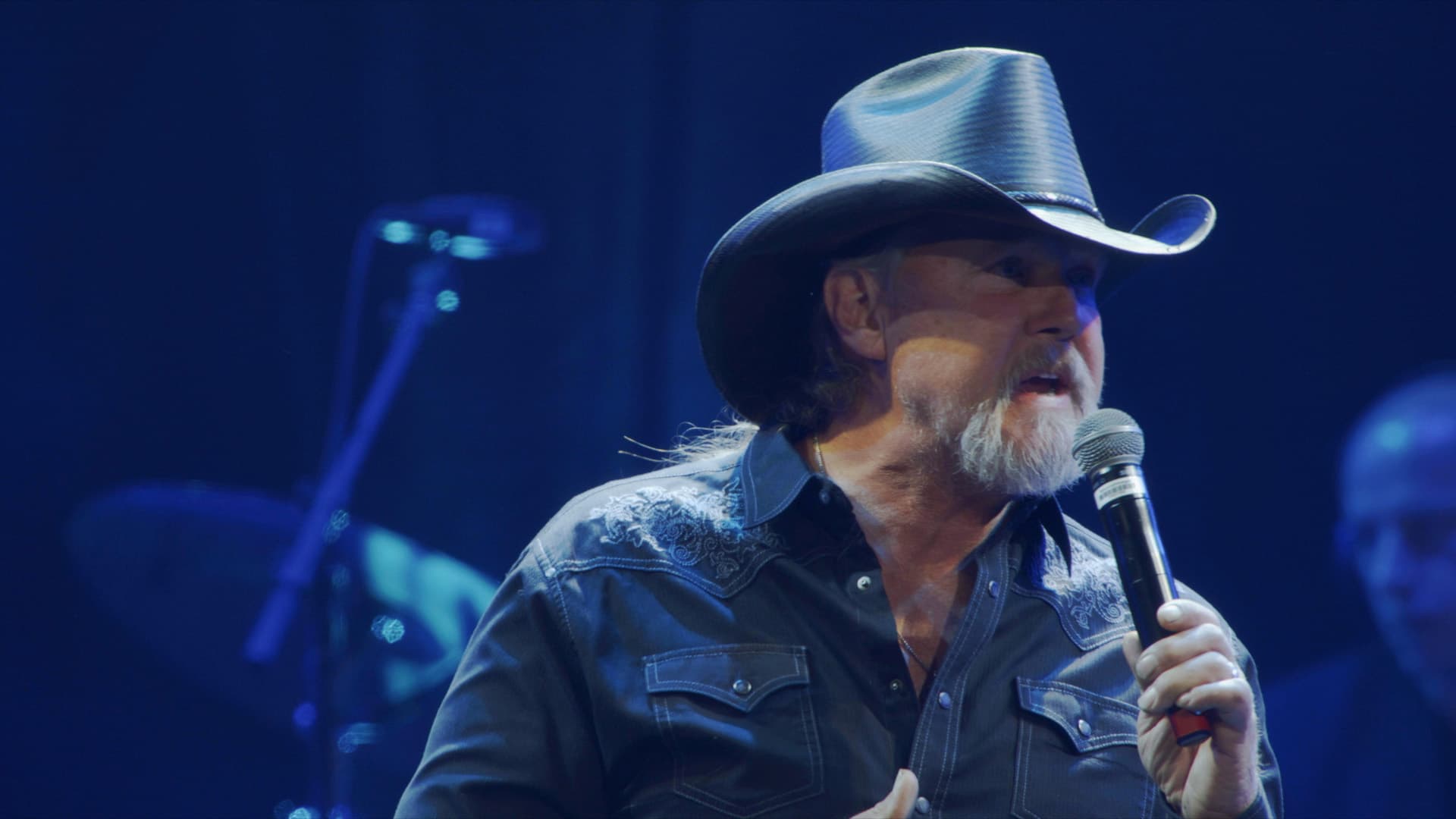 Trace Adkins in a black cowboy hat and shirt sings into a microphone on stage under blue lighting.