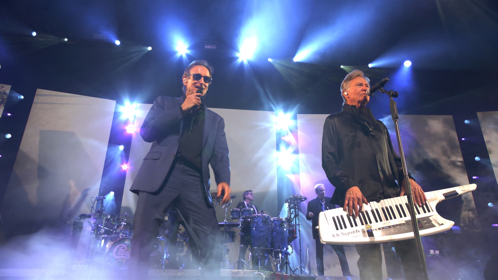 Chicago perform on stage, one with a microphone and the other holding a keytar, under bright stage lights.