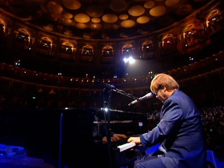 Elton John in a suit plays a grand piano and sings into a microphone on stage in a large, ornate concert hall filled with lights and an audience.