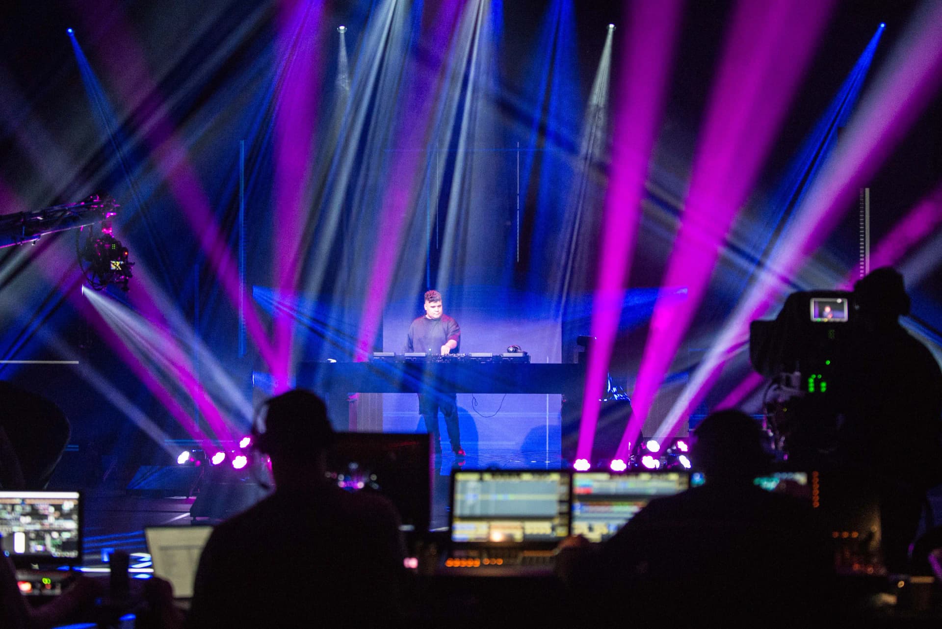 Justin Mylo performing on stage with vibrant pink and blue lights, surrounded by camera crews and equipment in the foreground.