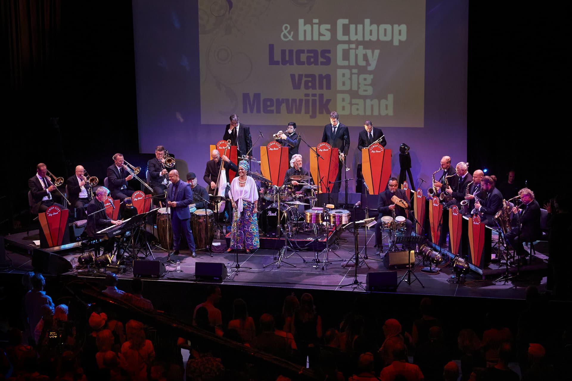 A large jazz big band performs on stage with multiple musicians playing brass, woodwind, and percussion instruments, backed by a projected sign reading “& his Cubop Lucas van Merwijk City Big Band.”.