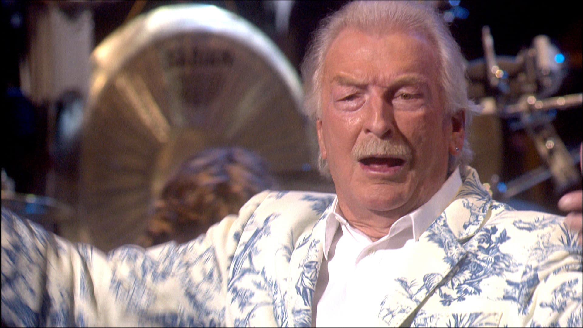 James Last in a floral jacket gestures energetically on stage, with musical instruments visible in the background.