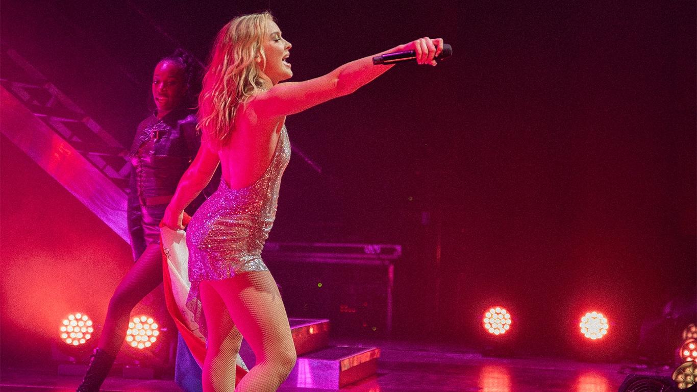 Zara Larsson singing on stage with a microphone, accompanied by a dancer in the background, under red and pink stage lights.