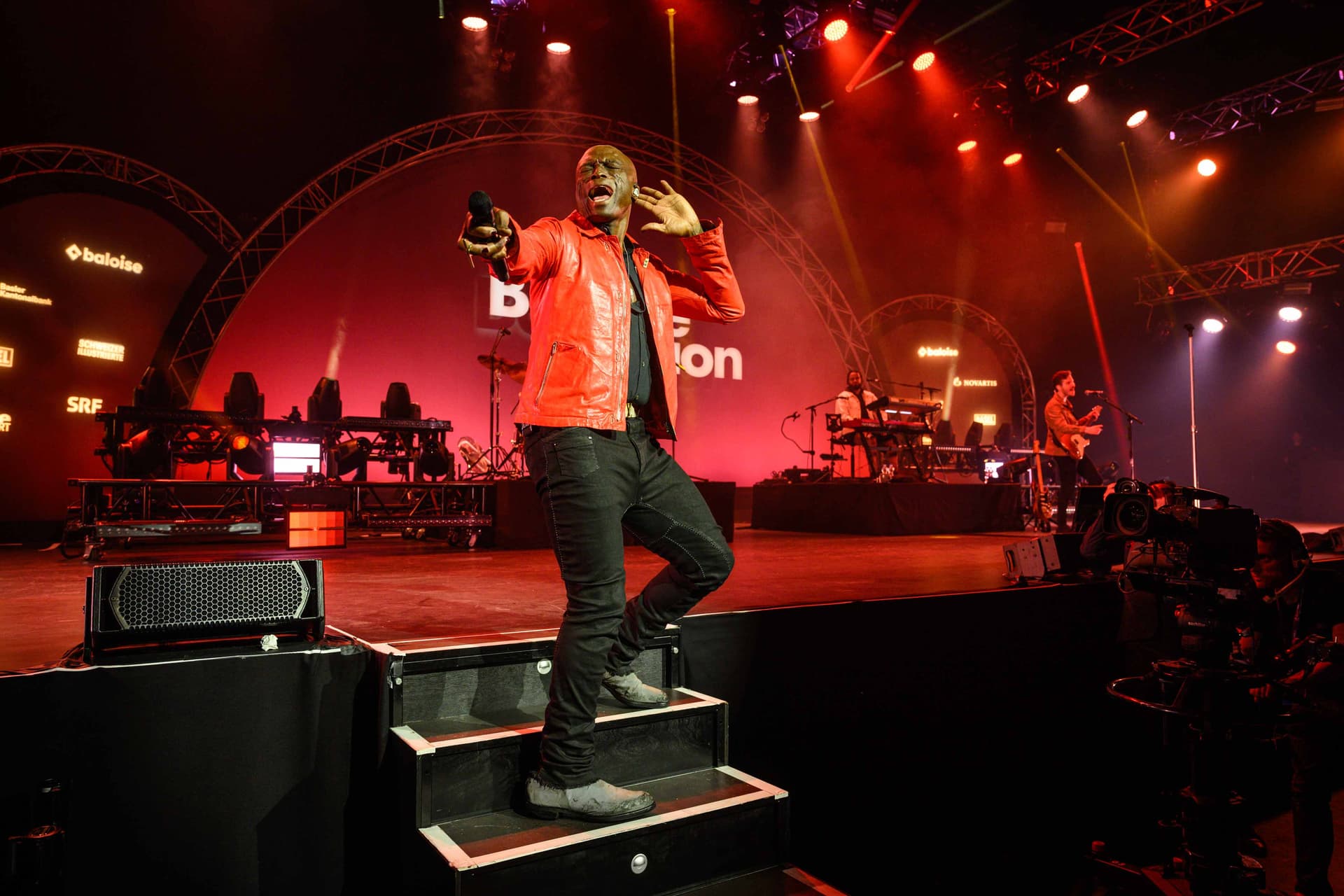 Seal in a red jacket sings on stage, holding a microphone, with band members and instruments in the background under colorful stage lights.