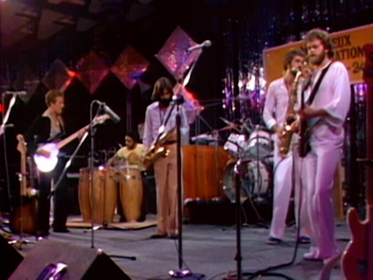 Average White Band performs on stage with instruments including a guitar, drums, congas, and saxophones. Multi-colored lights and a backdrop are visible.