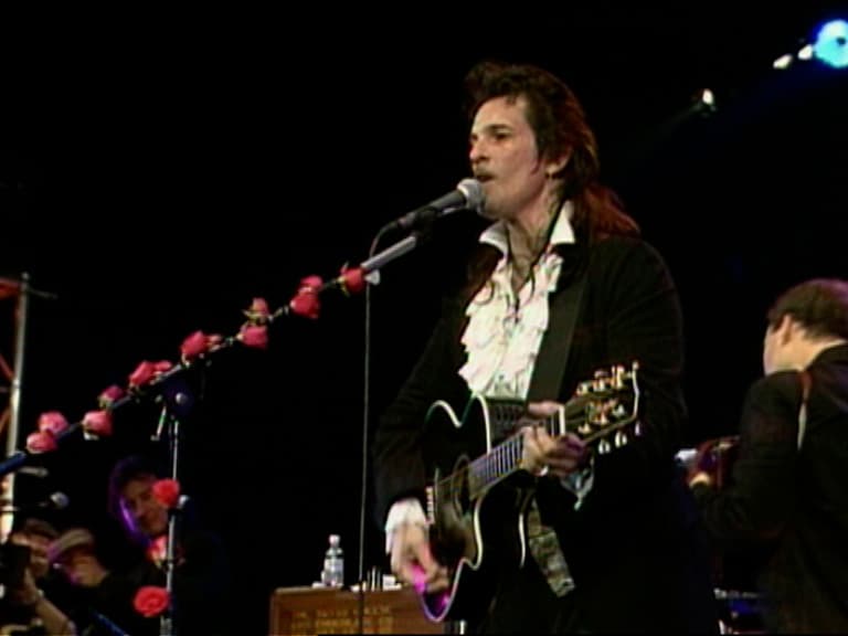Willy DeVille playing an acoustic guitar and singing into a microphone on stage, with red roses attached to the mic stand.