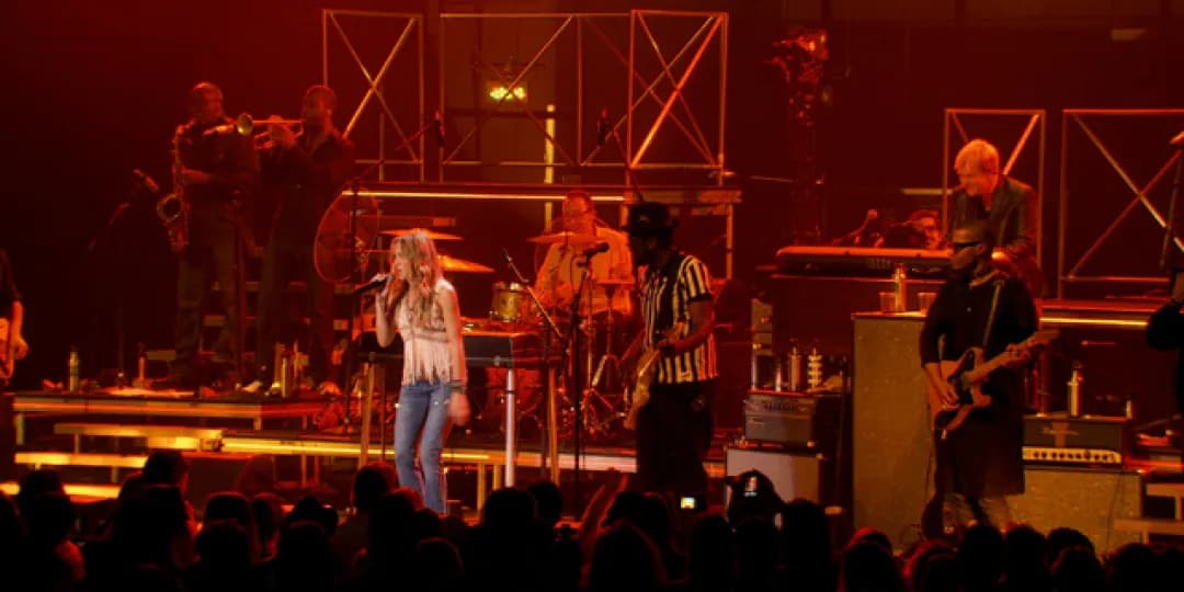 Sheryl Crow performing on stage under red lighting, featuring a lead singer, drummer, guitarists, and horn section. Audience visible in foreground.