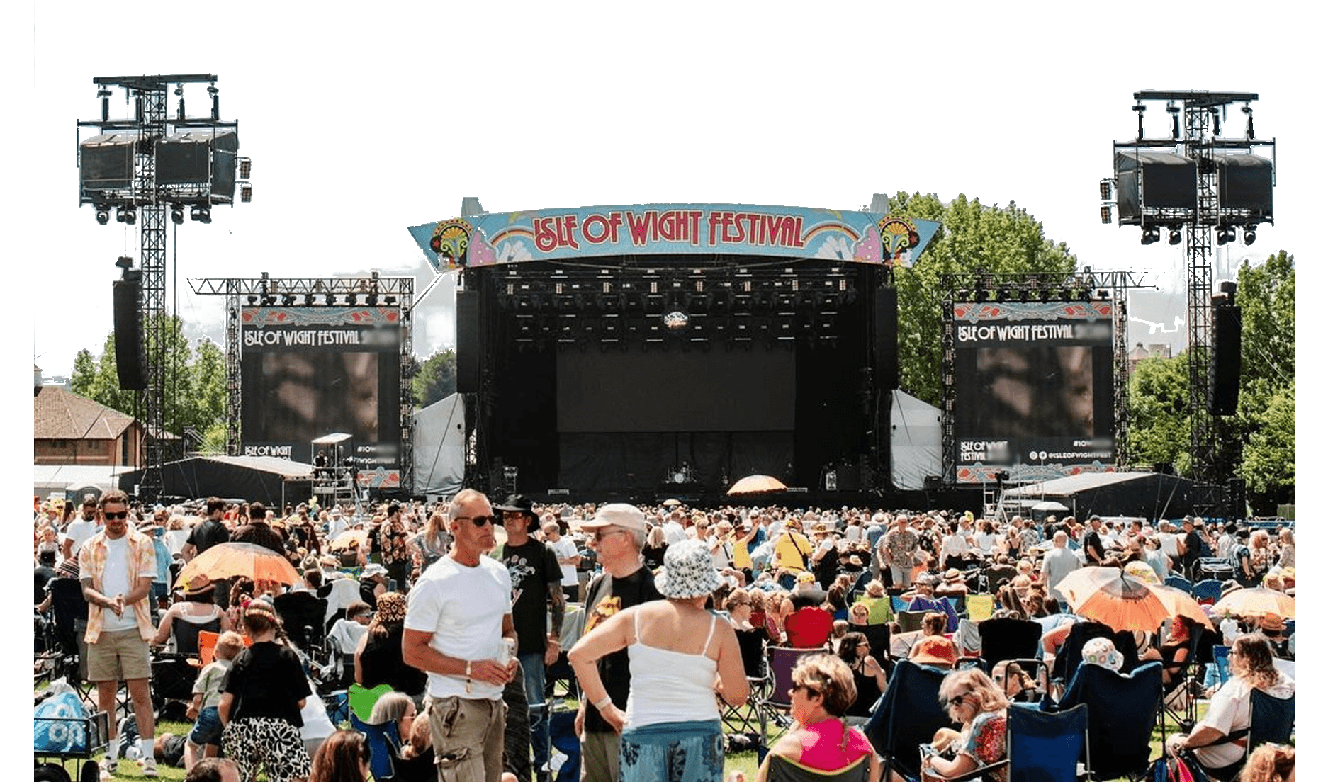 A large crowd sits and stands in front of the main stage at the Isle of Wight Festival during the day, with stage equipment and festival signage visible.