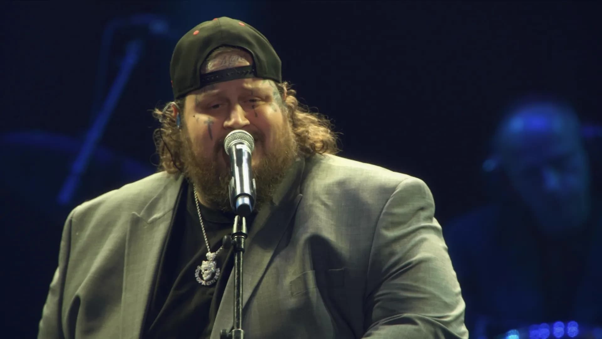 A man with a beard and long hair, wearing a backward cap, suit jacket, and necklace, sings into a microphone on stage.