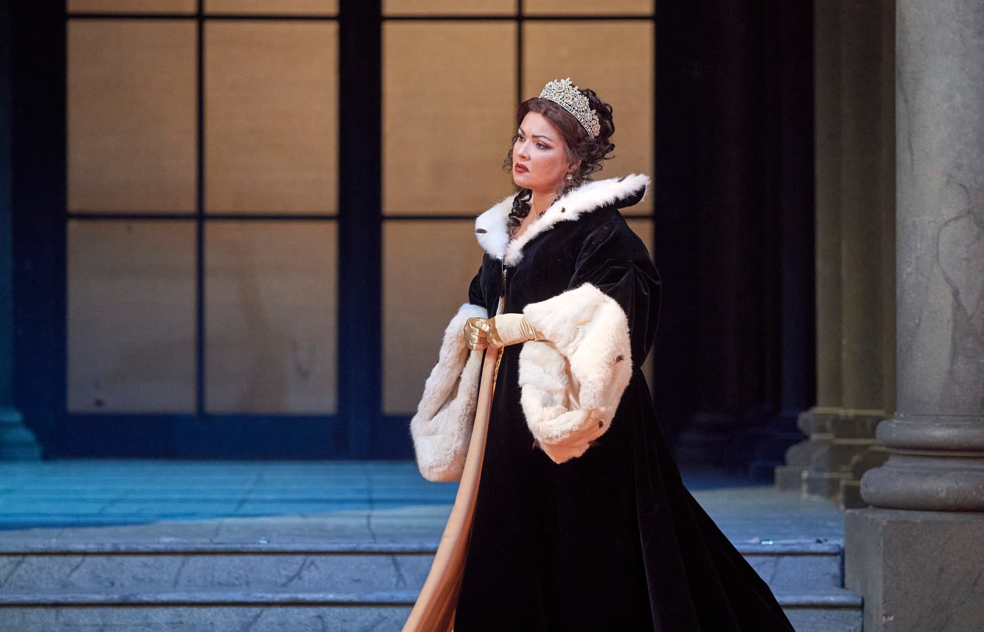 Taken from Tosca, a woman in an elaborate black and white fur-trimmed gown and tiara stands on a stone floor in front of tall windows and columns.