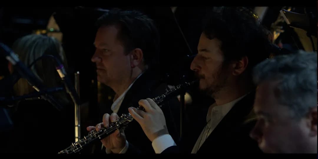 London Symphony Orchestra in formal attire perform in an orchestra pit. The focus is on a man playing the oboe, with other musicians and music stands visible nearby.