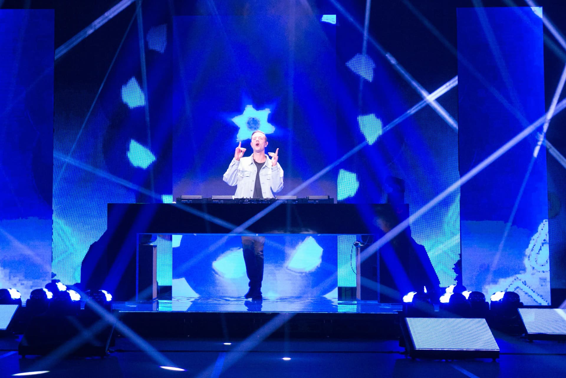 Sam Feldt performing on stage, surrounded by vibrant blue and white lights, standing behind a mixing console with a backdrop of abstract patterns.