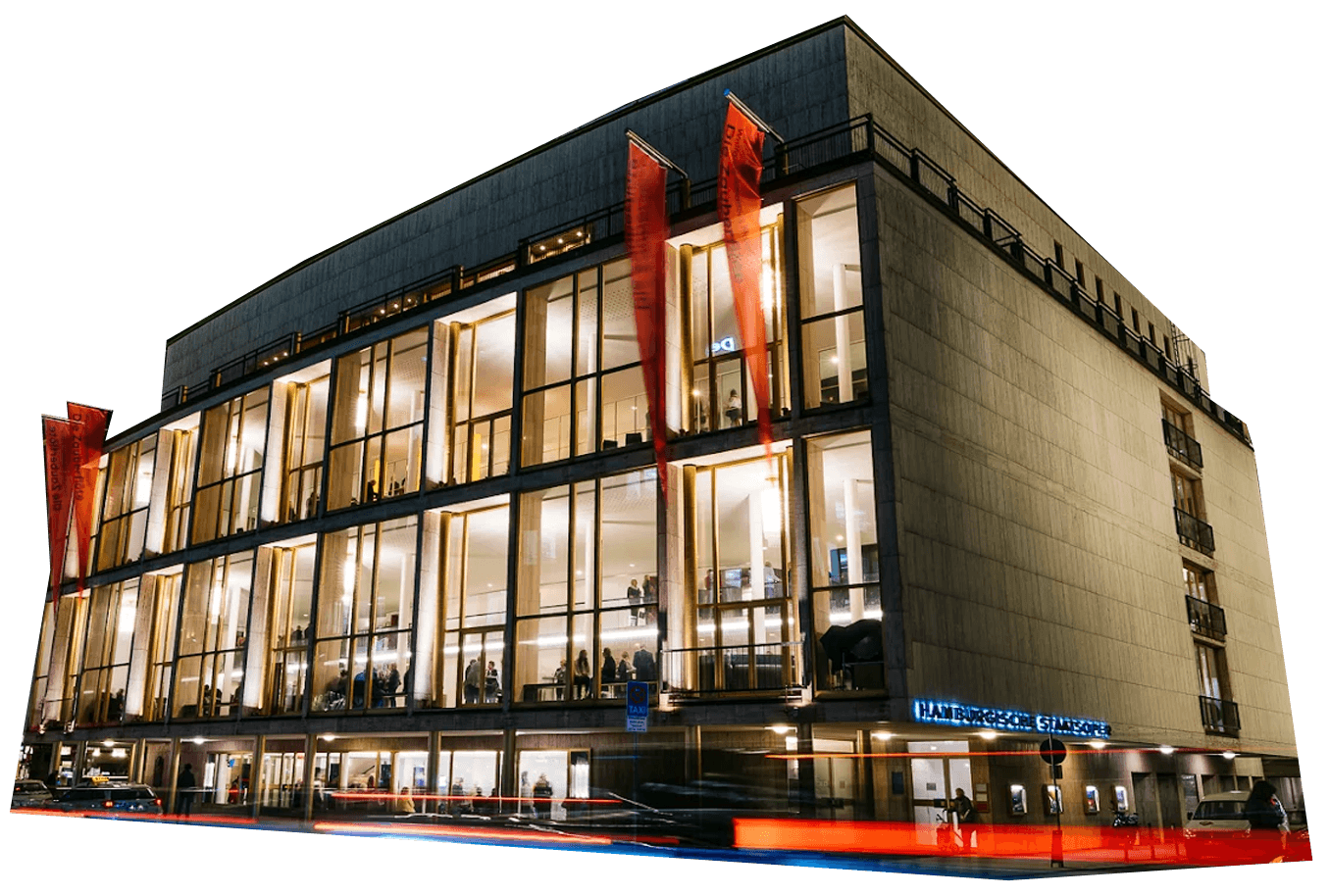 A modern, multi-story staatsoper hamburg building with large glass windows illuminated at night, displaying the sign "Hamburgische Staatsoper" above the entrance.