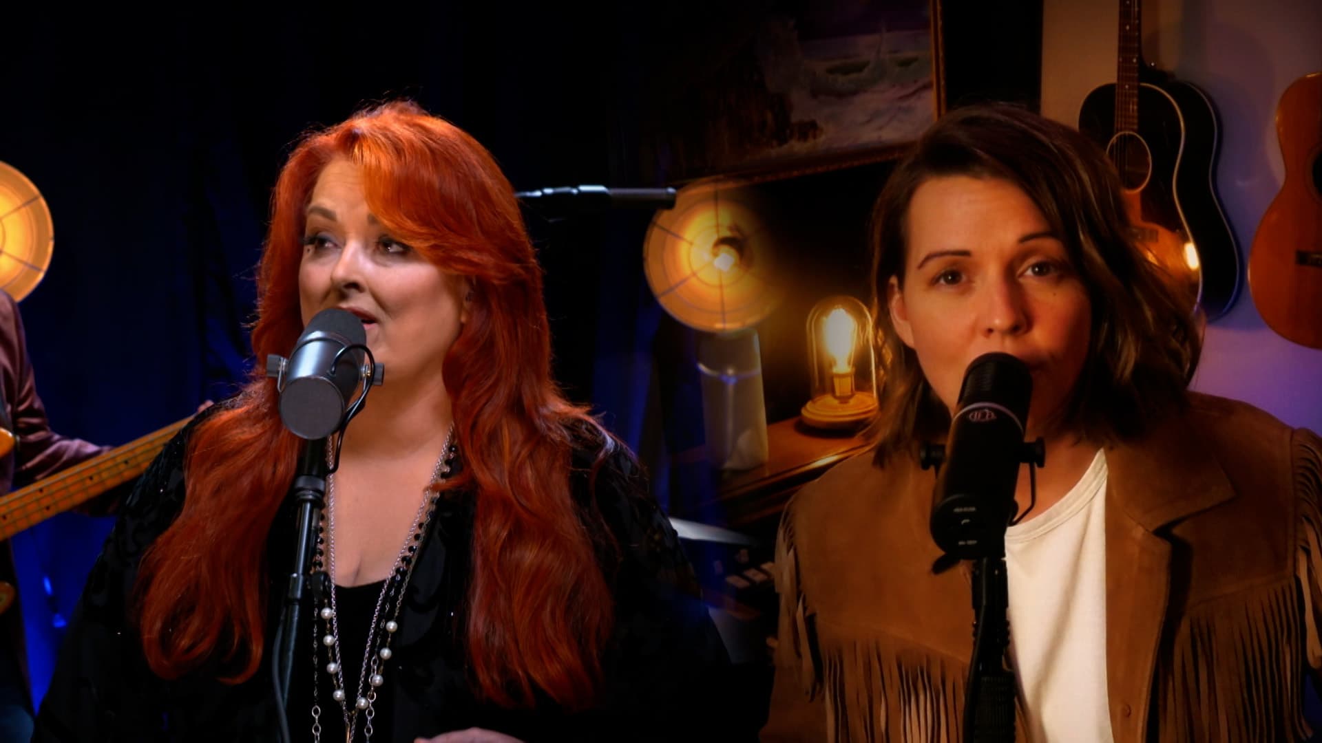 Two women sing into microphones on stage, one with long red hair and black attire, the other with short brown hair and a tan jacket; a lit lamp and paintings are visible in the background.