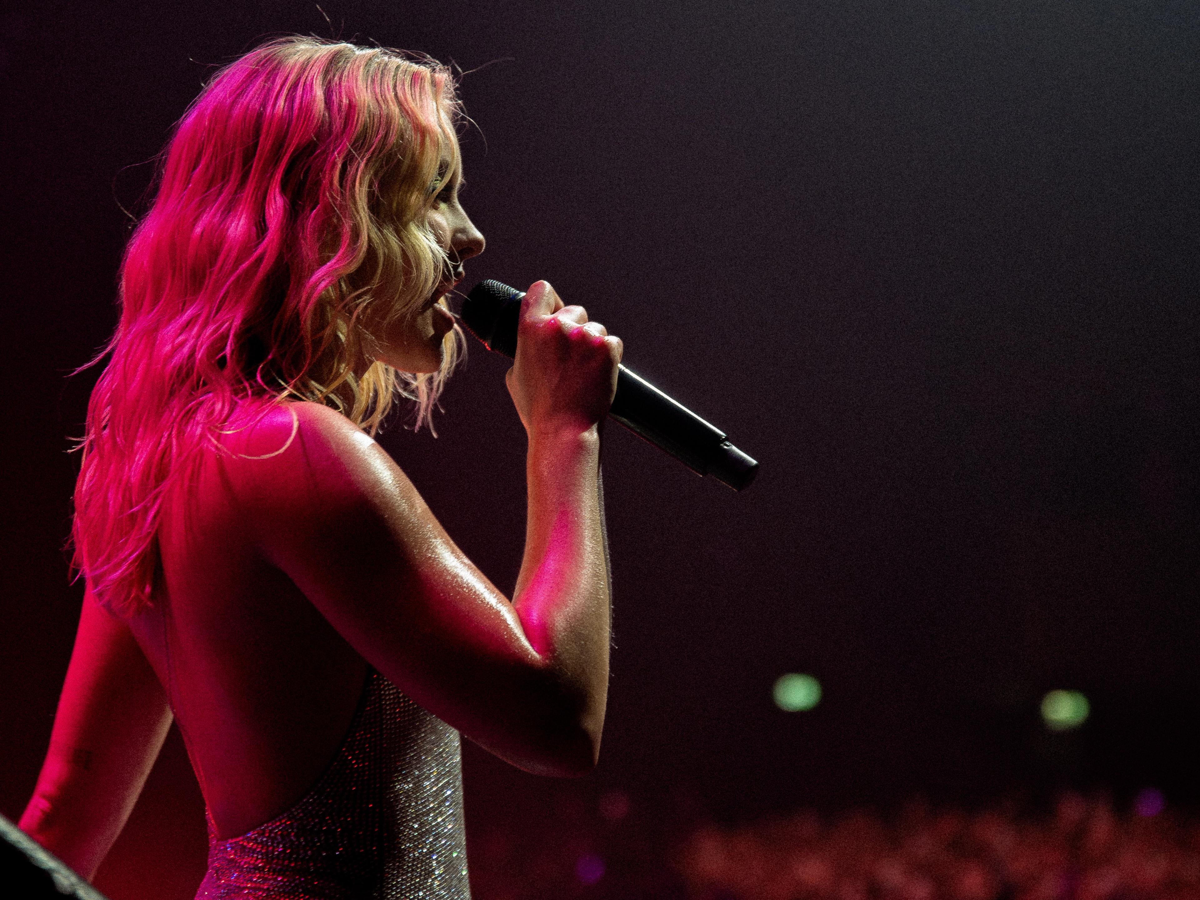 Zara Larsson singing on stage with a microphone in hand, illuminated by pink stage lights.