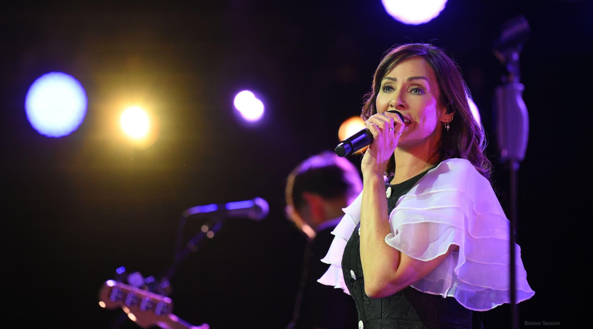 natalie imbruglia sings into a microphone on stage, wearing a black and white outfit, with colorful stage lights in the background and a guitarist playing beside her.