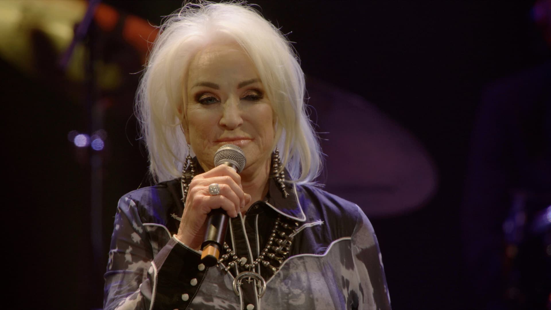 Tanya Tucker with white hair holds a microphone and performs on stage under concert lighting, wearing a dark patterned shirt and statement jewelry.