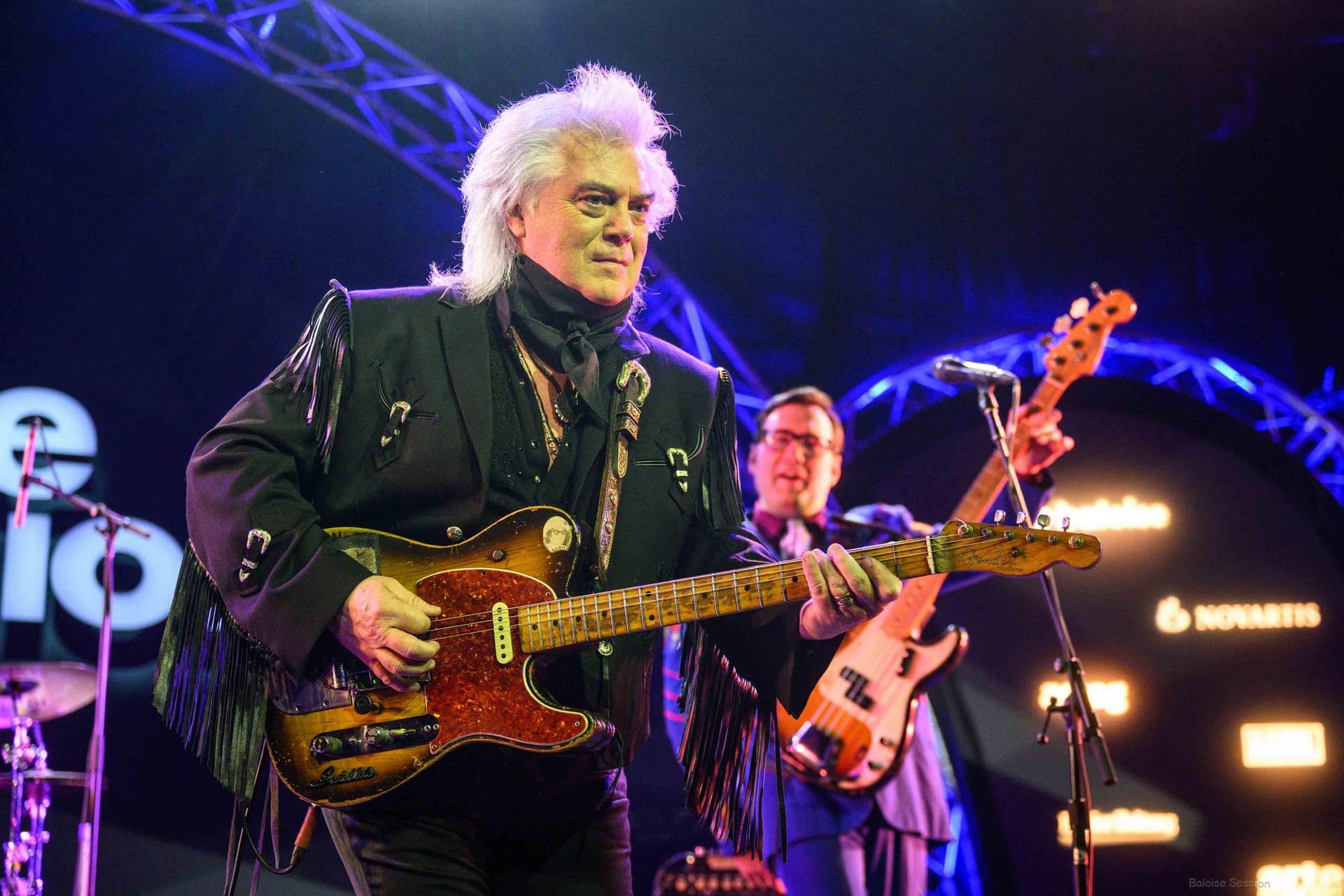 Marty Stuart with white hair plays an electric guitar on stage, wearing a black fringe jacket, with another band member playing bass guitar in the background.