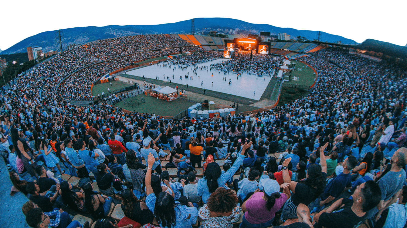 A large crowd fills a stadium, with people standing and sitting in the stands, while a stage with bright lights is set up on the field for a concert or event.
