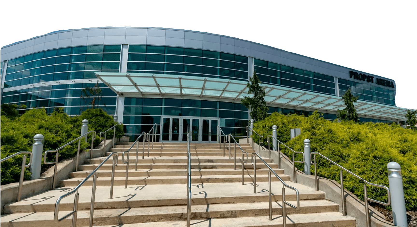 Wide concrete steps with metal railings lead up to the glass entrance of a modern building labeled "PROOST ARENA" under a blue sky.