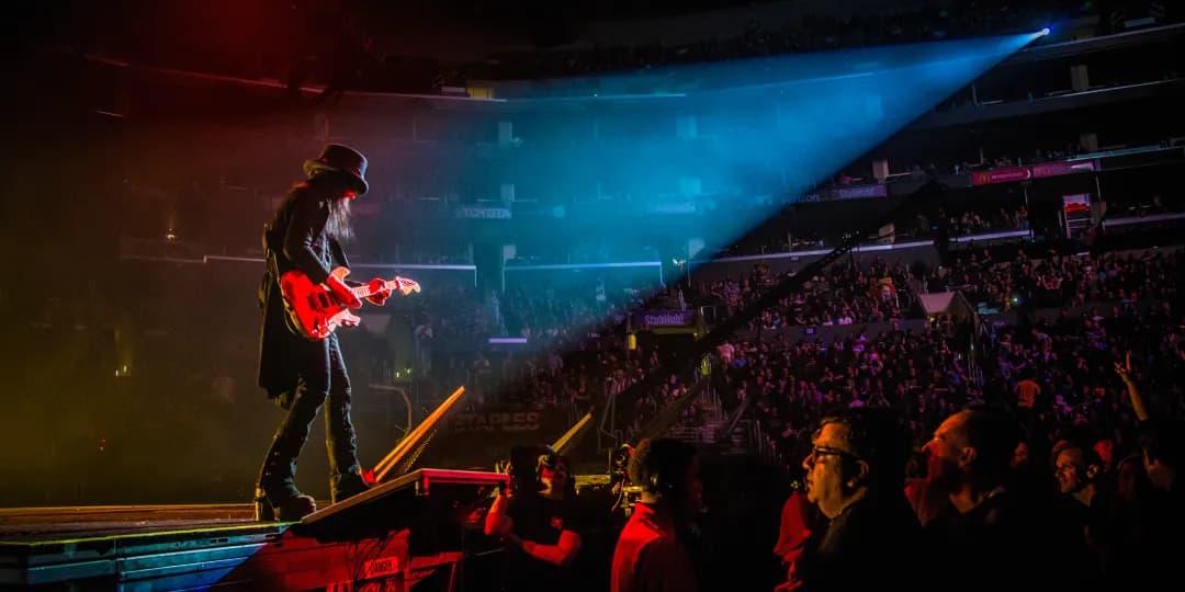 Motley Crue guitarist in a hat plays an electric guitar on a stage, illuminated by colorful lights, with a large audience in the background.