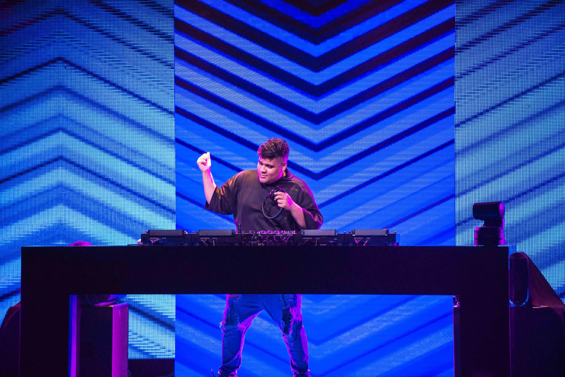 DJ Justin Mylo performing on stage with blue geometric pattern in the background, standing behind a mixing console, raising one hand.
