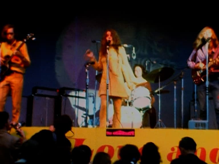 Big Brother & The Holding Company performs on an outdoor stage with a singer at the center, two guitarists on either side, and a drummer in the background. Audience members are visible in the foreground.