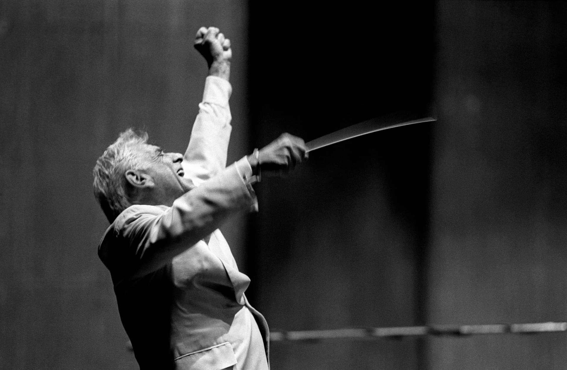 Leonard Bernstein in a suit raises one arm and holds a baton in the other, appearing to lead an orchestra with enthusiasm against a blurred background.