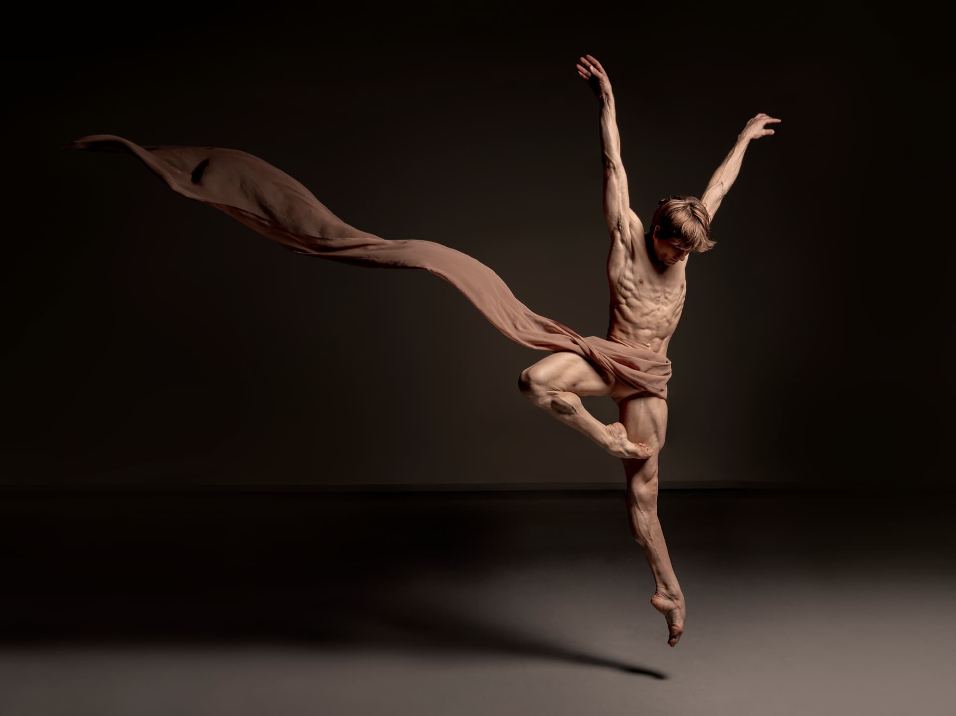 A male ballet dancer leaps mid-air with one leg raised and arms extended, wearing a flowing beige fabric against a dark background.