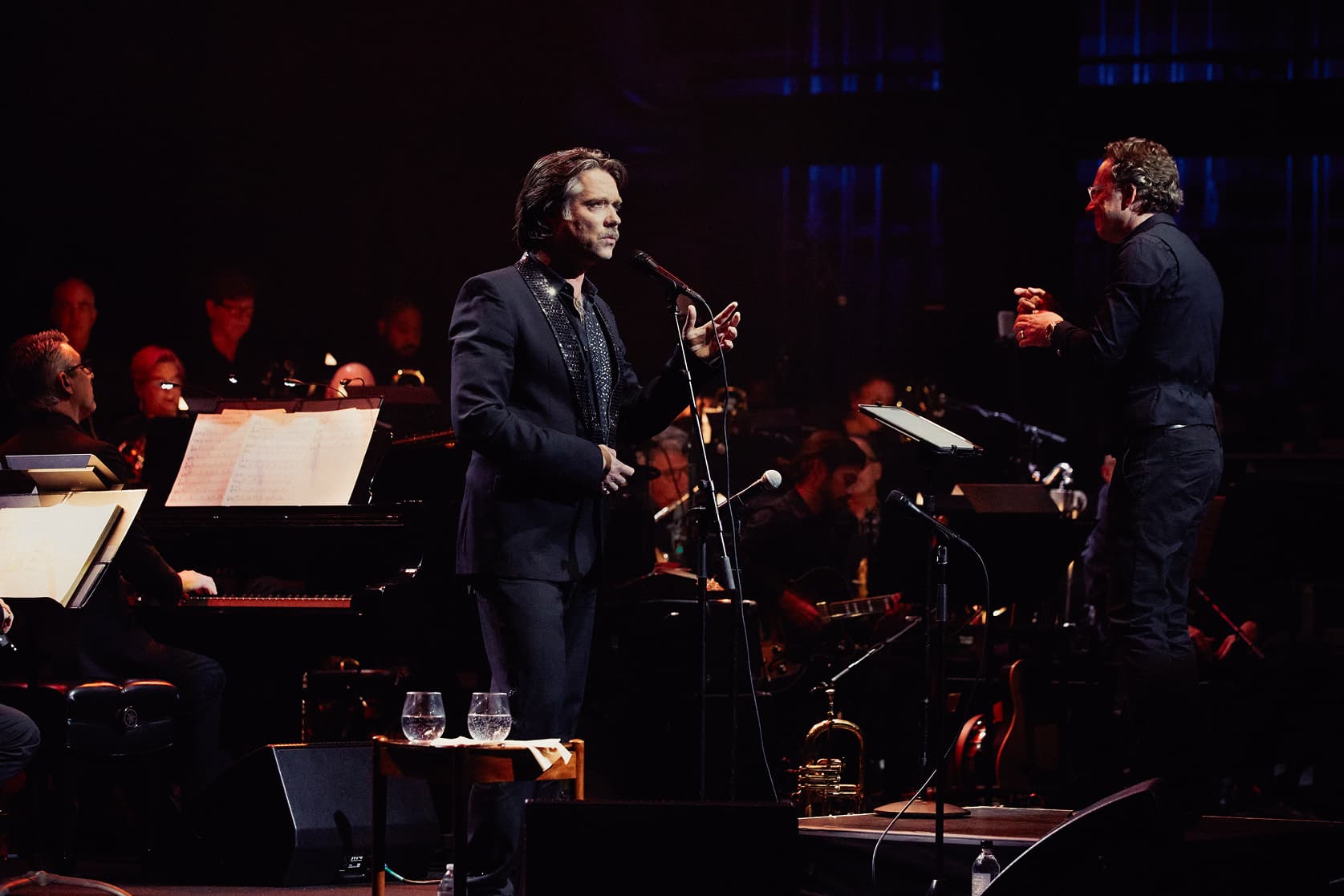 Rufus Wainwright in a black suit sings or speaks into a microphone on stage with a conductor and an orchestra playing behind him.
