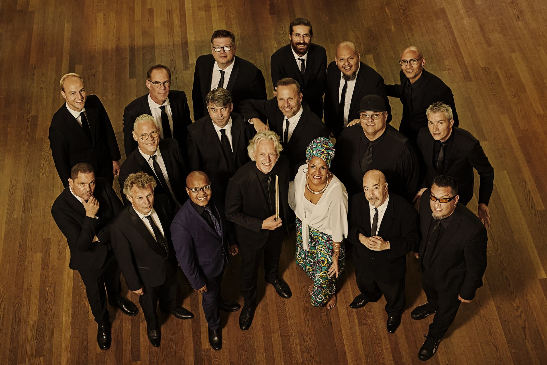 A group of nineteen adults dressed in formal attire stands in a semi-circle on a wooden floor, looking up at the camera.