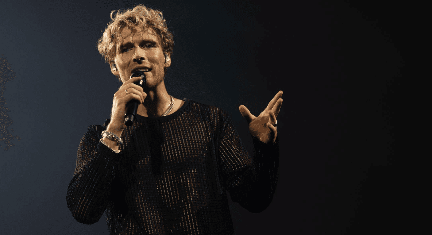 Christopher with curly blond hair sings into a microphone while wearing a black mesh shirt, standing against a dark background.