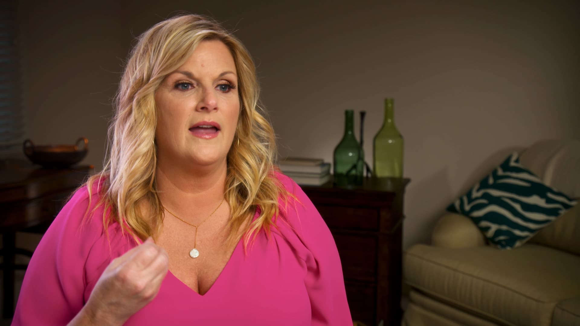 Trisha Yearwood with blonde hair wearing a pink blouse sits indoors, gesturing with her hand. There is a wooden table and bottles in the background.
