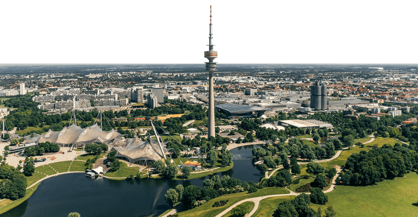 Aerial view of Munich’s Olympiapark with its distinctive tent-like stadium roof, a tall communications tower, surrounding buildings, and green park areas with water features.