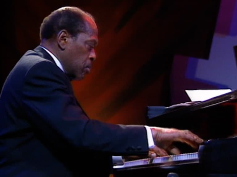 Kenny Drew in a dark suit plays a grand piano on stage, focusing on the keys. Warm lighting and abstract shapes are visible in the background.