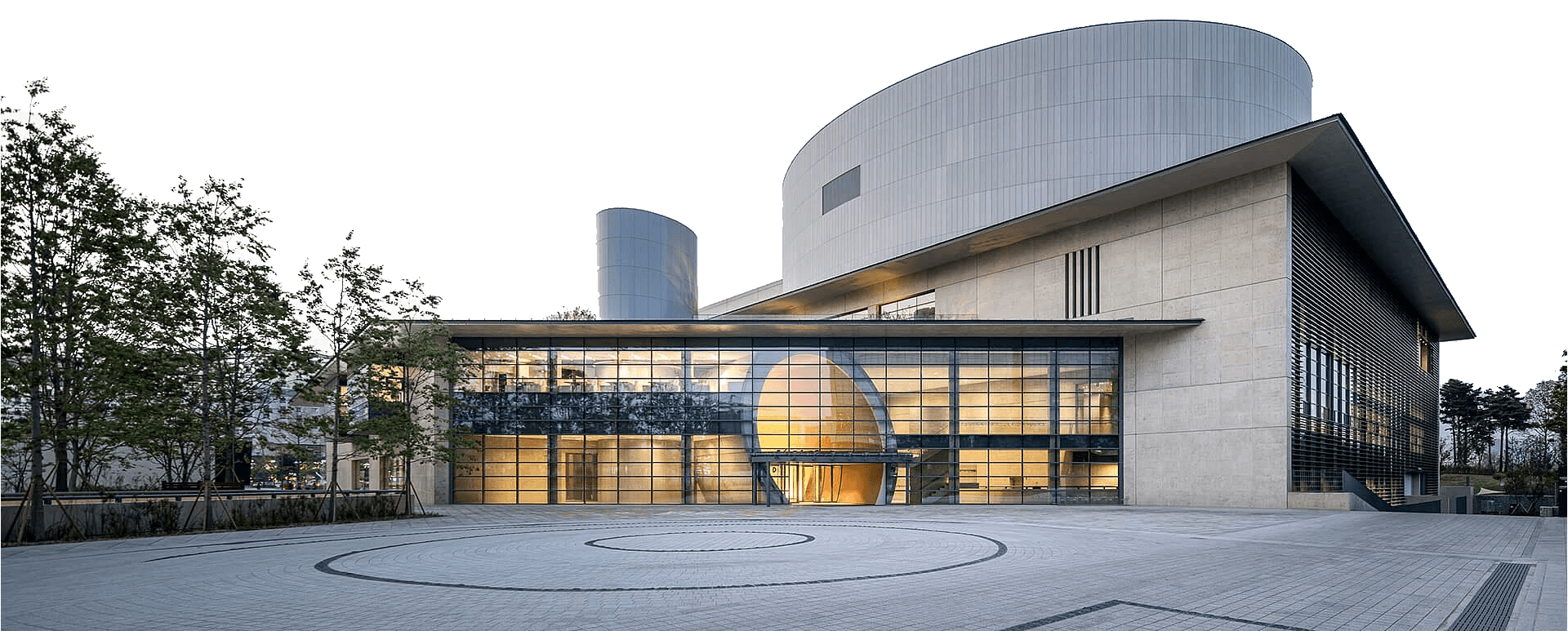 Modern LG arts center building with large glass windows, cylindrical structures, and a spacious paved courtyard with circular patterns; trees are visible on the left side.