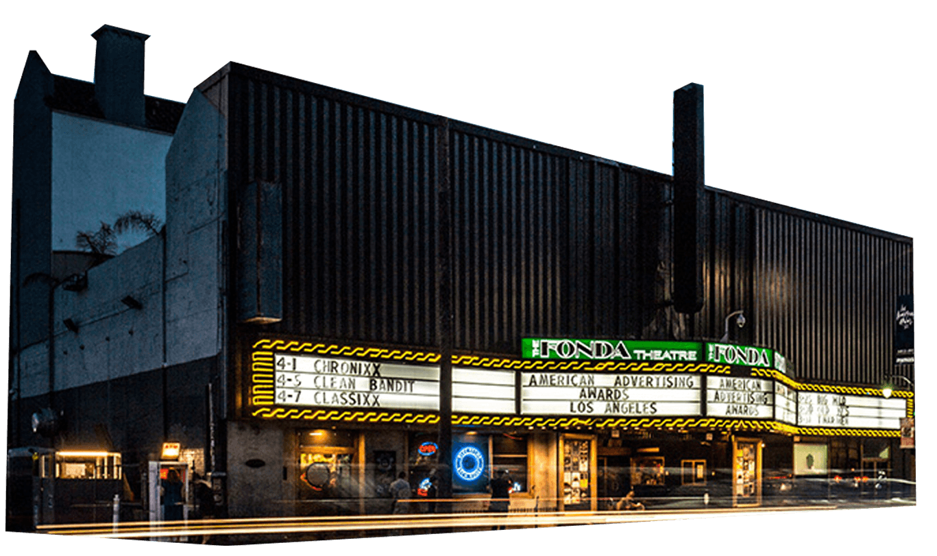 A street view of the Honda Theatre at dusk, with bright marquee signs advertising upcoming events and light streaks from passing cars.