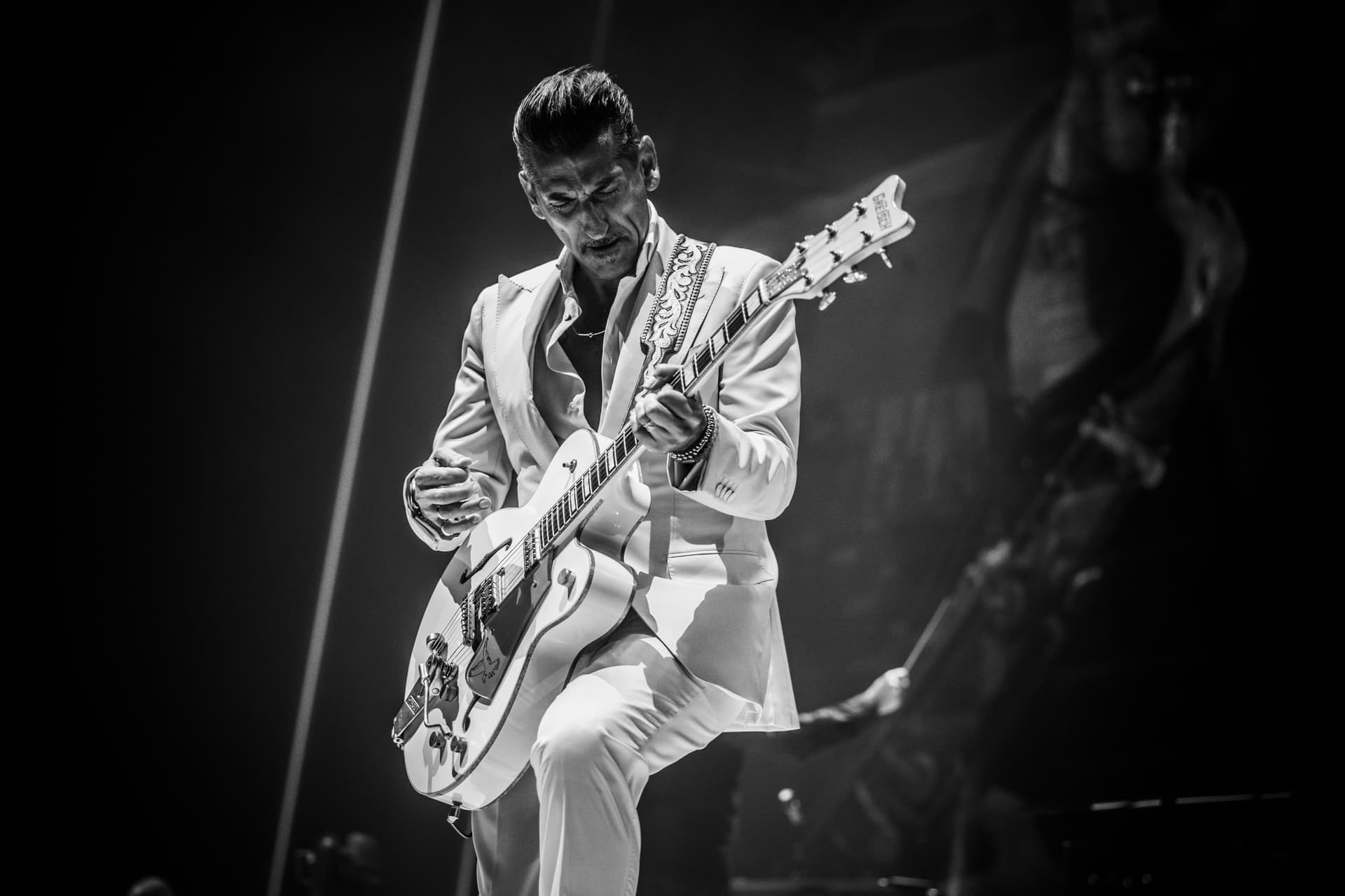 A man in a white suit plays an electric guitar on stage under dramatic lighting.