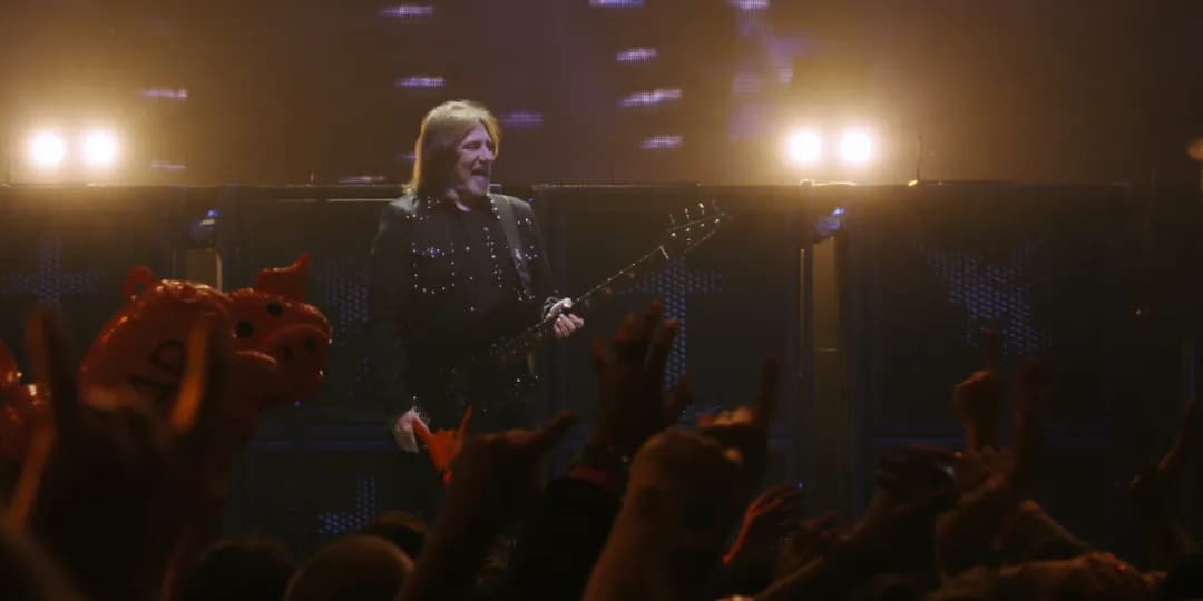 A musician playing bass guitar stands on stage under bright lights while an audience raises their hands and an inflatable dragon is visible in the crowd.