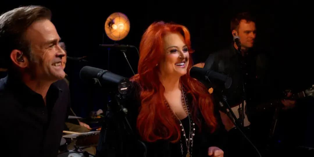 Three musicians perform on stage; a man sings at left, a woman with long red hair sings at center, and a guitarist plays at right with warm stage lighting behind them.