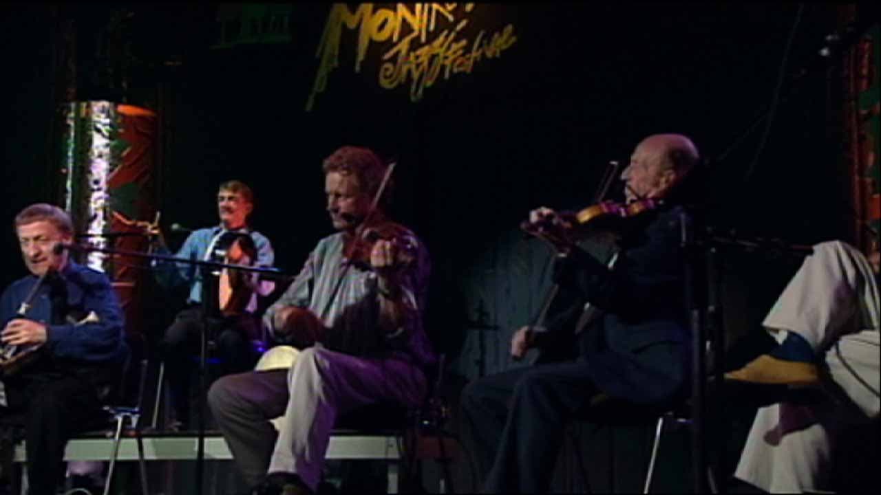 The Chieftans perform on stage, seated and playing traditional instruments including fiddles and pipes, with stage lighting and "Monday Night" signage in the background.