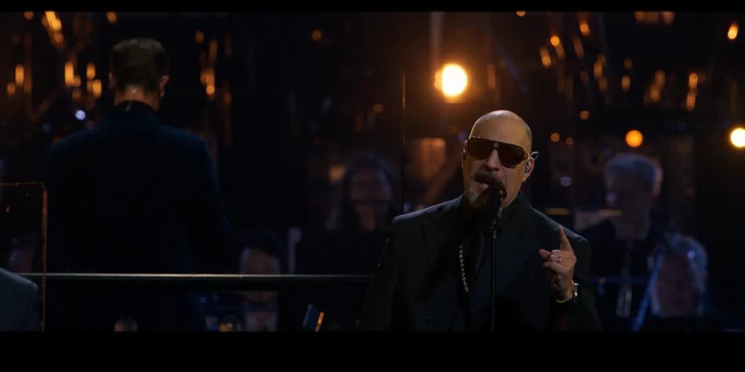 A man from Cypress Hill wearing sunglasses and a dark suit sings into a microphone on stage, with an orchestra and conductor in the background under stage lighting.