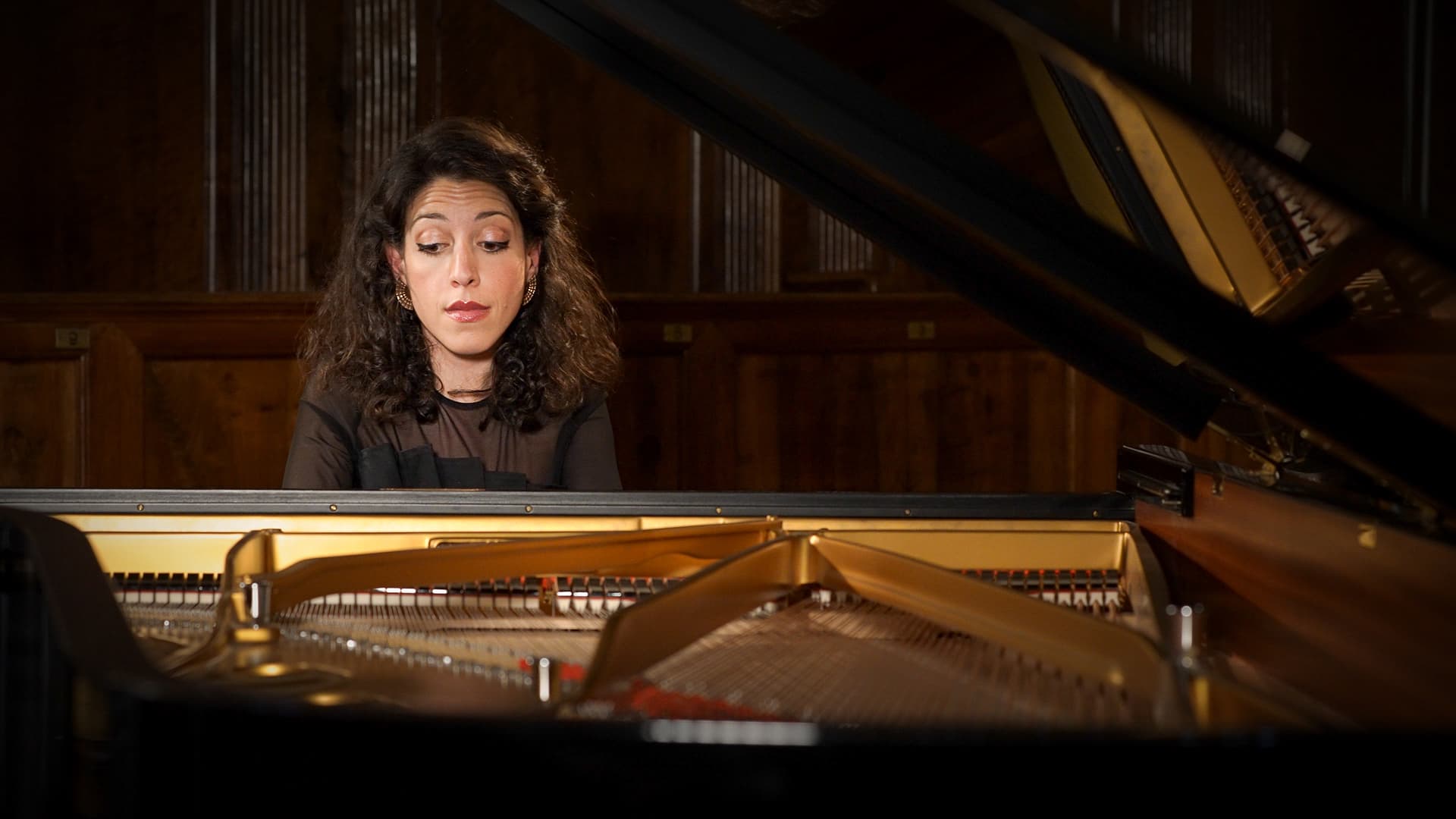Beatrice Rana with curly hair sits at a grand piano, appearing thoughtful while looking at the keys in a dimly lit room.