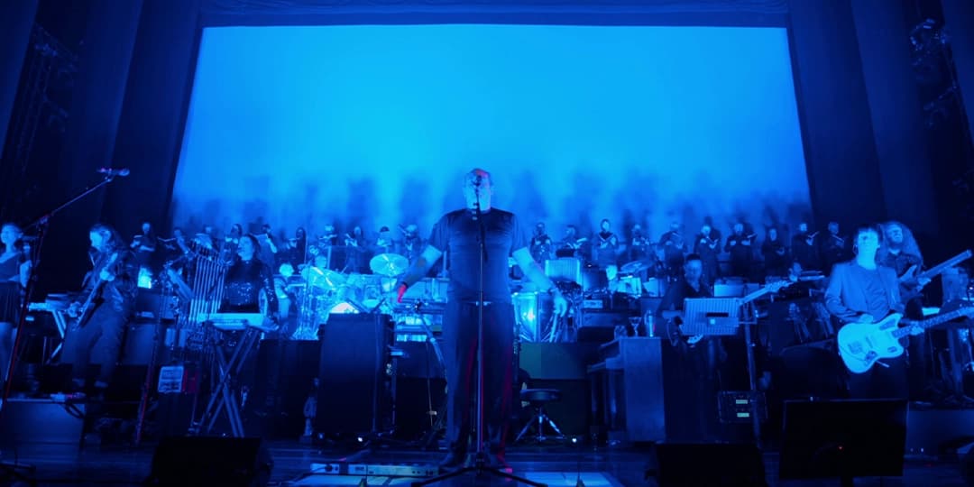 A Hans Zimmer live concert performance with a musician at center stage, surrounded by a band and a choir. The stage is illuminated with blue lighting.