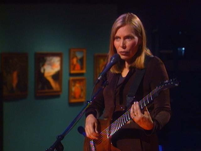 Joni Mitchell plays guitar and sings into a microphone on stage, with paintings in the background.