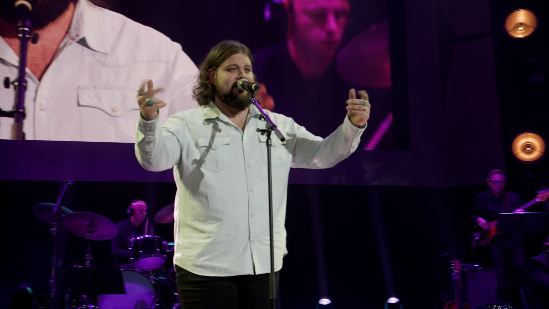 Dillon Carmichael sings into a microphone on stage with a band; a large screen behind him shows his image and a drummer.